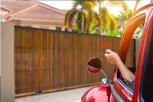 A person sitting inside a red car uses a remote control to open an electric gate leading to a modern house with a tiled roof. The surroundings include well-maintained greenery and palm trees in the background. The car's side mirror is visible.