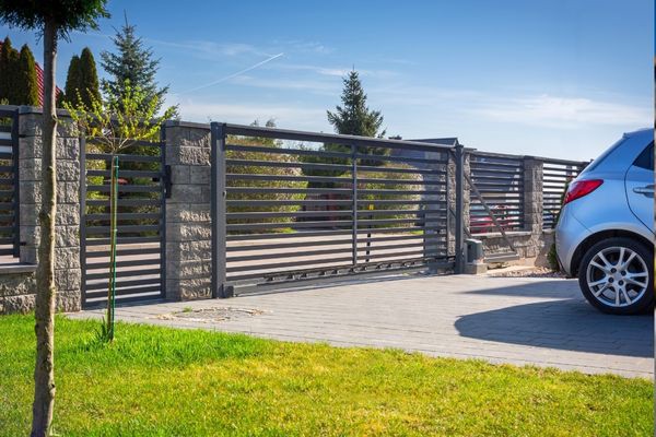 A silver car is parked next to a metal sliding gate with stone pillars. The electric gate leads to a driveway of a modern house with trees in the background. The scene is bright and clear under a sunny blue sky. A small tree and a green lawn are visible in the foreground.