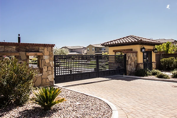 A sturdy, gated entrance to a residential community with a black, wrought-iron gate and stone pillars. The entrance is surrounded by well-manicured landscaping with shrubs and a clear, sunny sky overhead. Suburban homes are visible in the background, highlighting the expertise of local driveway gate repair technicians in San Antonio.