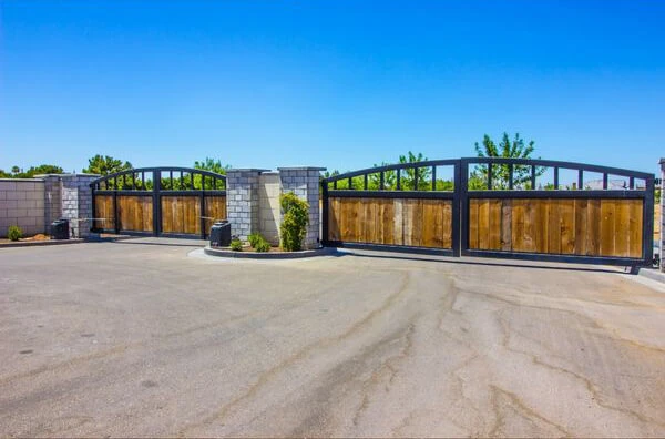 A wide metal entrance gate with wooden panels stands open, framed by stone pillars on a paved driveway. Green shrubs are planted at the base of the stone pillars, and trees are visible in the background under a clear, blue sky. The electric gate quietly awaits any necessary gate opener repair.