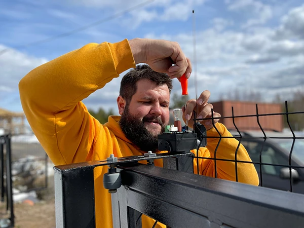 A bearded man in a yellow hoodie is using a screwdriver to install a mechanism on an electric gate with wire mesh fencing. The sky is partly cloudy, and the background features an outdoor landscape with trees and industrial buildings, showcasing the typical day of skilled gate technicians.