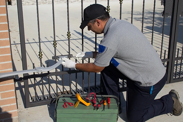 A man kneels on a concrete surface while performing driveway gate repair. He is wearing a cap, a gray shirt, dark pants, and white gloves. In front of him is an open toolbox with various tools scattered around it. The black gate has vertical metal bars, highlighting the meticulous task at hand.