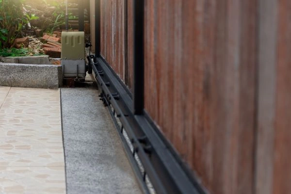 Close-up view of a sliding gate mechanism next to a stone-paved path. The gate features black metal tracks and wooden panels, with some foliage visible in the background. The morning sunlight gently illuminates the scene from the top left corner, showcasing excellent craftsmanship by San Antonio's gate technicians.