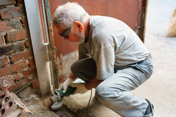 An older man with gray hair and beard, wearing a gray T-shirt and pants, is crouched down using a power tool to cut or grind a metal surface at a construction site. Safety glasses protect his eyes as he works on what appears to be an electric gate repair beside a brick wall and metal structure.