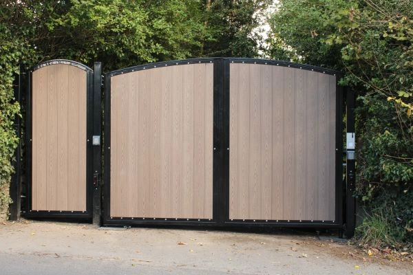 Large, dual-panel wooden gate with a smaller pedestrian gate on the left, framed in black metal. This beautiful electric gate is closed, surrounded by lush green foliage and set against a concrete or paved surface. For maintenance or repairs, skilled gate technicians in San Antonio are always available.