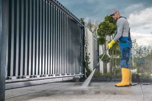 A person wearing a cap, safety goggles, gloves, overalls, and yellow boots is using a power washer to clean an outdoor tiled area next to a fence on a cloudy day, perhaps preparing for gate technicians in San Antonio.