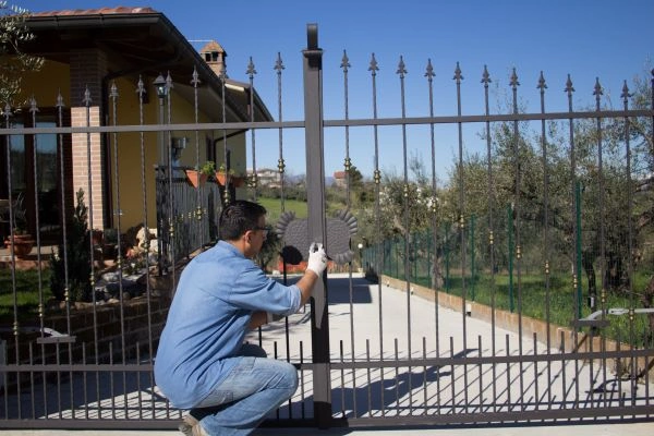 A man wearing a blue shirt and jeans is crouching and installing or repairing a metallic gate. He is using tools and working outdoors in a residence with a long driveway in San Antonio, with a brick house visible in the background. The sky is clear and blue.