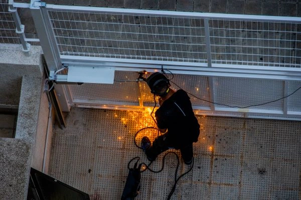 A person wearing protective gear welds metal at the base of a white metal structure, possibly preparing for an electric gate installation. Sparks and bright lights are visible from the welding process, and tools are scattered around on the ground. The image is taken from an overhead angle.