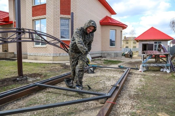 A person in camouflage clothing works on the construction of a metal framework for an electric gate in an outdoor area near a brick house with red roofing. They are using tools to adjust metal beams, surrounded by construction materials and equipment typically used by gate technicians.