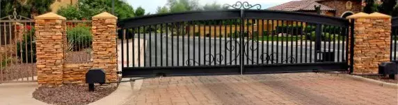 An electric wrought iron gate spans the entrance to a driveway, flanked by stone pillars and fencing. The gate is set against a backdrop of a landscaped property with trees and a house visible in the distance. The brick-paved driveway leads to this elegant entrance, maintained by skilled gate technicians.