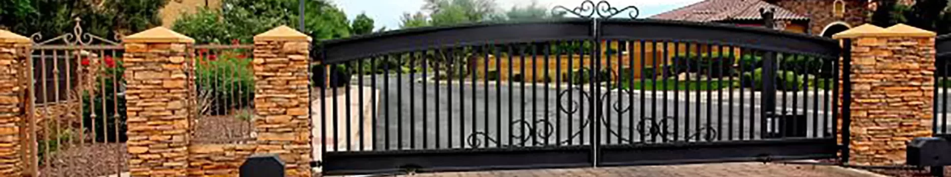 A tall, wrought-iron gate stands closed, flanked by two stone columns. Behind the gate, a wide driveway leads to a house partially visible on the right. Trees and bushes line the driveway, and the sky is clear. An electric gate system ensures smooth access and enhances security for residents.